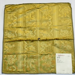 May include: A gold-colored fabric swatch featuring a damask pattern of floral motifs and ribbons. A white label in the bottom right corner displays the text Scalamandre, OAK HILL, Damask, Color: Gold, 100% Silk, 51 inch Approx Width, 11 inch Approx Vert Rpt.