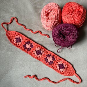 May include: A handmade crochet headband in coral with square floral designs in deep purple. Three balls of yarn in shades of pink and purple are also visible, suggesting a crafting theme. The headband is designed with a long, thin strap.