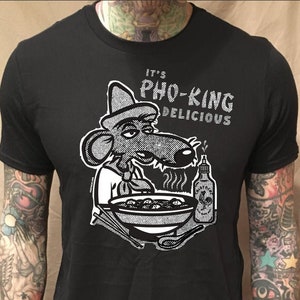 May include: Black t-shirt with a white graphic of a cartoon rat wearing a hat and eating pho. The text "It's Pho-King Delicious" is printed on the shirt.