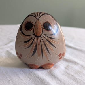 May include: A rounded ceramic owl figurine featuring a smooth, light tan surface with hand-painted black and reddish-brown details. The design includes large circular eyes, a pointed beak, radiating feather patterns, and small three-dot clusters.