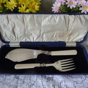 Puede incluir: Una caja de terciopelo azul con forro de satén blanco, que contiene un cuchillo de mantequilla y un tenedor de plata con mangos de marfil.