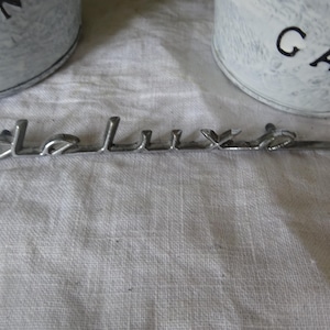 Puede incluir: Un emblema de coche de metal plateado con la palabra "de luxe" en relieve.