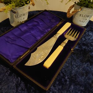 Puede incluir: Un juego antiguo de cuchillo y tenedor de plata con mangos color marfil, exhibido en una caja forrada de terciopelo. Los utensilios de plata tienen diseños grabados ornamentados. La caja está abierta, revelando el interior de terciopelo morado.