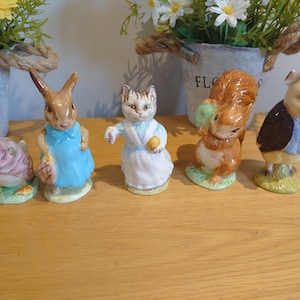 May include: A set of ceramic figurines depicting characters from a children's story. The figurines include a duck, rabbit, cat, squirrel, and pig, each with detailed clothing and accessories. The figurines are set on a wooden surface.