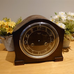 Könnte beinhalten: Eine dunkelbraune hölzerne Kaminuhr mit rundem Zifferblatt und goldfarbenen Ziffern. Die Uhr wird von kleinen Metalleimern mit gelben und weißen Blumen flankiert. Das Zifferblatt hat eine silberfarbene Umrandung.
