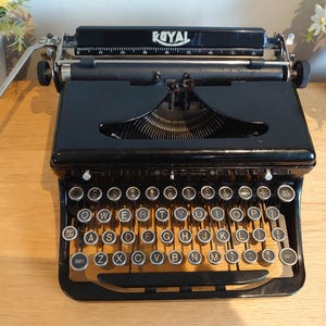 Könnte beinhalten: Eine schwarze Royal Schreibmaschine mit QWERTY-Tastaturlayout und runden Tasten, einem metallenen Zeilenschalthebel auf der linken Seite und einer sichtbaren Papierwalze. Der Markenname Royal ist in Weiß auf dem oberen Gehäuse aufgedruckt.