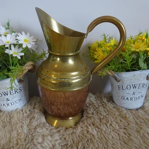 Può includere: Una brocca decorativa in ottone e rame con un manico curvo e un beccuccio largo. La brocca è affiancata da due piccoli secchi bianchi con le parole "FLOWERS & GARDEN" e pieni di fiori bianchi e gialli.