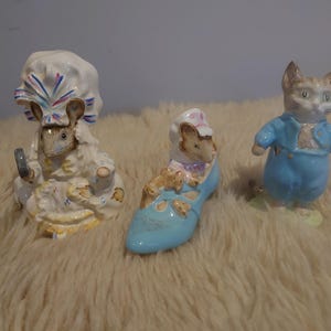 Puede incluir: Tres figuras de cerámica: un ratón con un gorro, un ratón en un zapato azul y un gato con una chaqueta azul. Las figuras de ratón llevan ropa blanca y amarilla. La figura del gato lleva una chaqueta y pantalones azules.