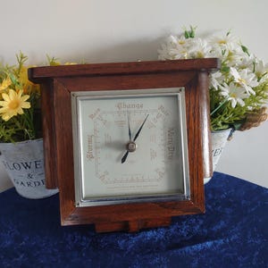 May include: A square wooden barometer with a dark brown frame and a white face. The face features black text including Stormy, Rain, Change, Fair, and Very Dry, with two black hands pointing toward the center. The device has a silver metal border.
