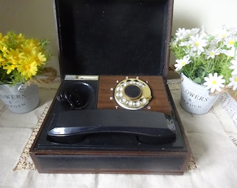 May include: A vintage rotary dial phone in a wooden case with a black receiver and a gold-colored dial. The phone is on a white tablecloth with a floral pattern.
