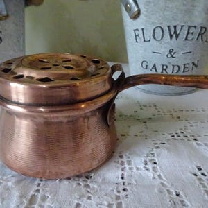 Puede incluir: Una pequeña olla de color cobre con una tapa con múltiples agujeros pequeños. La olla tiene un asa y está sobre un mantel de encaje blanco. Dos pequeños cubos de metal con las palabras "FLOWERS & GARDEN" están en el fondo.