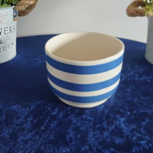 May include: A small, round ceramic bowl with a cream-colored interior and exterior. The exterior features horizontal blue and white stripes. The bowl is sitting on a blue surface. Two small decorative flower pots are in the background.