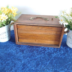 May include: A vintage wooden box with three drawers, a brass handle, and a keyhole. The box is surrounded by two small galvanized metal buckets filled with yellow and white flowers. The buckets have the words "FLOWERS & GARDEN" printed on them.