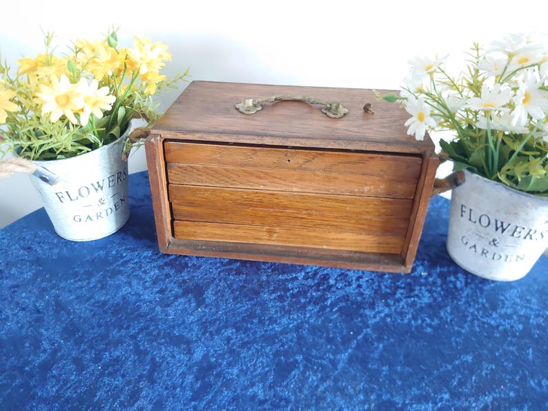 May include: A vintage wooden box with three drawers, a brass handle, and a keyhole. The box is surrounded by two small galvanized metal buckets filled with yellow and white flowers. The buckets have the words "FLOWERS & GARDEN" printed on them.