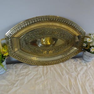 May include: A decorative brass tray with intricate filigree patterns and handles. The oval tray features a central well and is flanked by small potted flowers. A unique piece for serving or display.