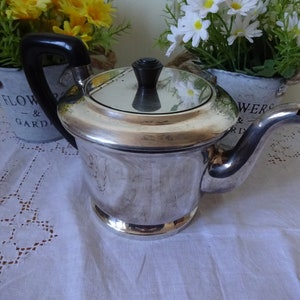 Puede incluir: Una tetera de plata con un asa y un pomo de la tapa de plástico negro. La tetera tiene una superficie brillante y reflectante y un cuerpo redondo. Está sentada sobre un mantel blanco con un patrón de encaje.