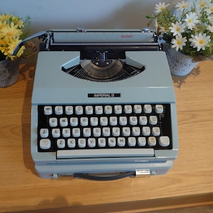 May include: A vintage light blue Imperial II typewriter with white keys and black lettering. The word "SIGNET" is visible on the top. The typewriter sits on a wooden surface, with potted flowers in the background.