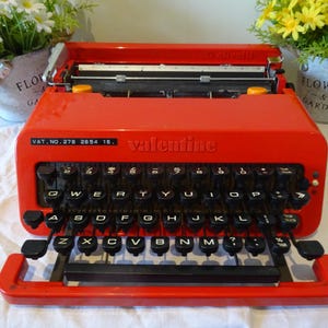 Peut inclure: Une machine à écrire Valentine rouge vif avec des touches noires. Le mot "valentine" est imprimé sur le devant. La machine à écrire a une disposition de clavier QWERTY standard. Le texte "VAT. NO.278 2854 15." est également visible.