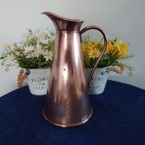 May include: A copper-coloured jug with a curved handle and spout. The jug is in front of two small white flower pots with rope handles, each filled with flowers. The pots are labelled "Flower & Garden".