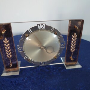 May include: An Art Deco clock with a silver face and black numerals. The clock is flanked by two rectangular panels with gold wheat stalk designs on a dark blue background. The clock is supported by a clear base.