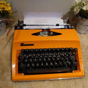 May include: A bright orange vintage typewriter with black keys and paper inserted. The typewriter sits on a fluffy, light-coloured surface. Two small metal buckets with flowers are on either side of the typewriter.