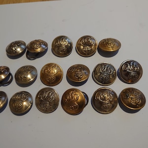 Może przedstawiać: Kolekcja antycznych mosiężnych guzików z ozdobnymi wytłoczonymi wzorami. Guziki mają różne kolory, od złotego do brązowego, a niektóre mają pętelki z tyłu. Guziki ułożone są na jasnoszarej powierzchni.