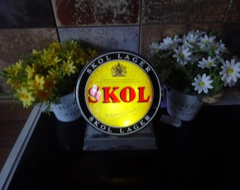 May include: A round, black and yellow sign with the words "Skol Lager" printed around the edge. The center of the sign features the word "Skol" in red letters on a yellow background.