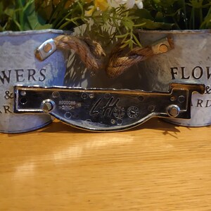 Puede incluir: Dos macetas de metal galvanizado con asas de cuerda y las palabras "FLOWERS & GARDEN" impresas. Una placa de metal brillante con el texto "LH" y otras marcas est&aacute; delante de las macetas.