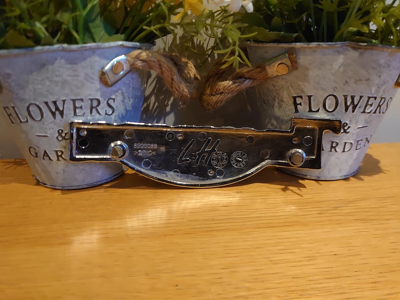 Puede incluir: Dos macetas de metal galvanizado con asas de cuerda y las palabras "FLOWERS & GARDEN" impresas. Una placa de metal brillante con el texto "LH" y otras marcas est&aacute; delante de las macetas.