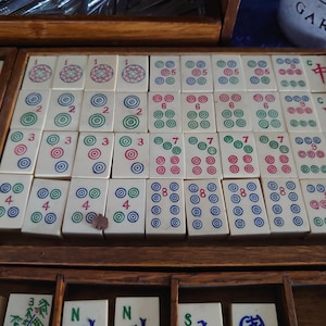May include: A wooden tray filled with ivory-colored Mahjong tiles. The tiles display various combinations of circles and Chinese characters in red, green, and blue. The tiles are arranged in rows and columns within the tray.