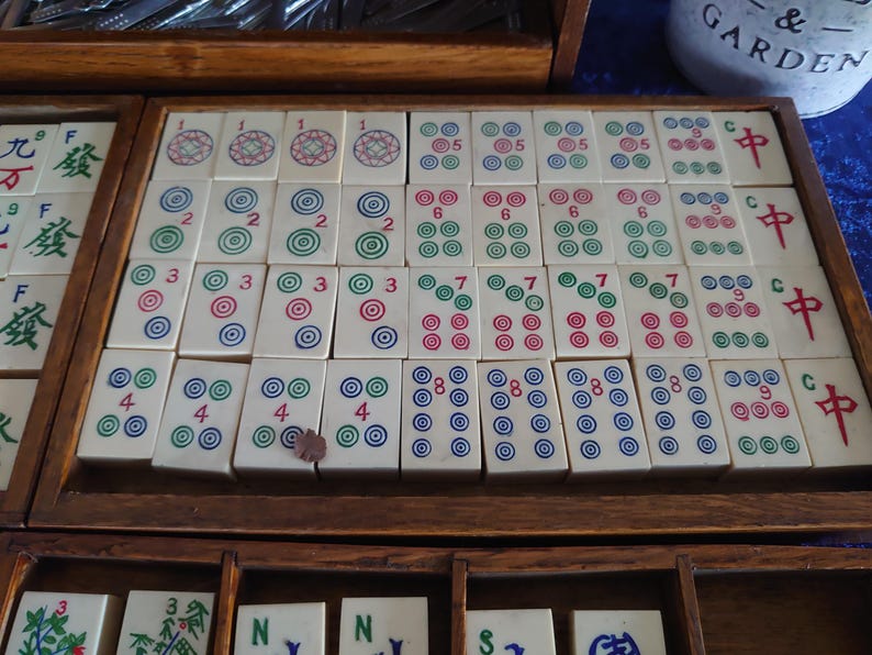 May include: A wooden tray filled with ivory-colored Mahjong tiles. The tiles display various combinations of circles and Chinese characters in red, green, and blue. The tiles are arranged in rows and columns within the tray.