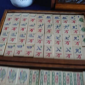 May include: A vintage Mahjong set with ivory-colored tiles, each displaying Chinese characters and symbols in red, blue, and green. The tiles are arranged in wooden trays, showcasing the game's components.