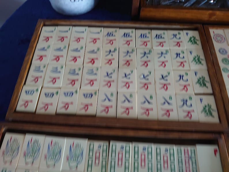 May include: A vintage Mahjong set with ivory-colored tiles, each displaying Chinese characters and symbols in red, blue, and green. The tiles are arranged in wooden trays, showcasing the game's components.