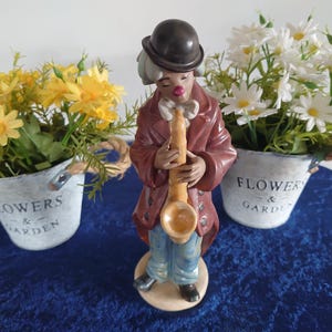 May include: A ceramic clown figurine wearing a dark brown coat, blue trousers, and a black bowler hat, playing a tan saxophone. The clown has a white ruffled collar and a small red nose, standing on a circular base between two metal buckets.