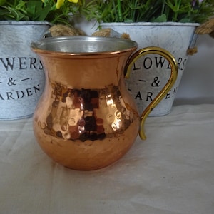 Puede incluir: Una taza de color cobre, con acabado martillado y asa dorada. El interior es plateado. La taza está delante de dos macetas de metal blanco con las palabras "FLOWERS & GARDEN" impresas.