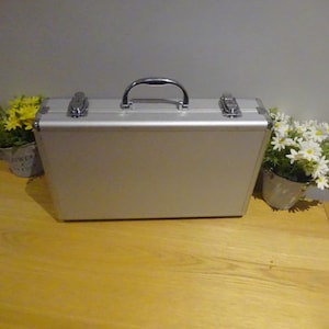 Puede incluir: Maletín plateado con asa negra y dos cierres plateados. El maletín está sobre una superficie de madera, con pequeñas plantas en macetas con flores amarillas y blancas a ambos lados.