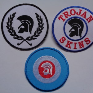 May include: Three embroidered patches with a black and white image of a helmet with a laurel wreath, the words "TROJAN SKINS" in red and blue, and a red and white helmet on a blue and white circle.