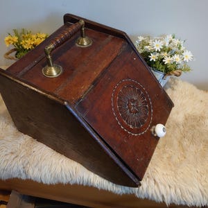 Peut inclure: Une boîte en bois brun foncé avec une poignée et des détails en laiton. La boîte présente un motif circulaire sculpté et un bouton blanc en céramique. La boîte est inclinée et repose sur une surface moelleuse de couleur crème.