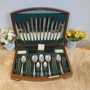 Könnte beinhalten: Eine Holzkiste mit einem Besteckset, ausgekleidet mit grünem Samt, gefüllt mit silbernen Utensilien. Die Kiste ist geöffnet und zeigt Messer, Gabeln und Löffel. Zwei kleine Blumentöpfe mit weißen und gelben Blumen.
