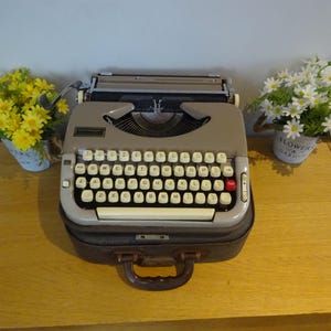 May include: A vintage typewriter with a gray body and white keys, resting on a brown case with a handle. The typewriter is flanked by small pots of yellow and white flowers, creating a classic aesthetic.