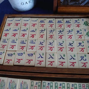 May include: A wooden tray filled with ivory-colored Mahjong tiles. The tiles feature Chinese characters and numbers in red, blue, and green. The tray is part of a larger set, with additional tiles visible in the background.