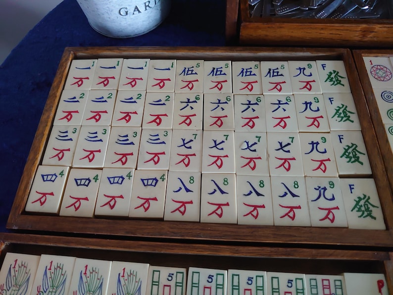 May include: A wooden tray filled with ivory-colored Mahjong tiles. The tiles feature Chinese characters and numbers in red, blue, and green. The tray is part of a larger set, with additional tiles visible in the background.