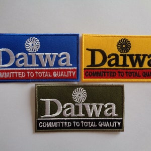 Può includere: Tre toppe ricamate con il logo Daiwa. Le toppe sono blu, gialle e verdi. Ogni toppa presenta il testo "Daiwa" in lettere bianche o nere e il testo "Committed to Total Quality" in lettere rosse.