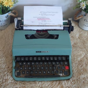 May include: A teal Olivetti Lettera 32 typewriter with a QWERTY keyboard layout, black keys, and a red margin release key. A sheet of white paper is inserted into the carriage, featuring typed text including the words Made in England.