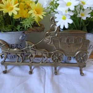 Könnte beinhalten: Ein Schlüsselhalter aus Messing in Form einer von Pferden gezogenen Kutsche mit drei Haken. Die Kutsche ist detailliert mit einem Kutscher und Fenstern. Der Schlüsselhalter wird auf einer weißen Oberfläche mit zwei weißen Blumentöpfen mit den Aufschriften "Flowers Garden" und "Flowers" gezeigt.