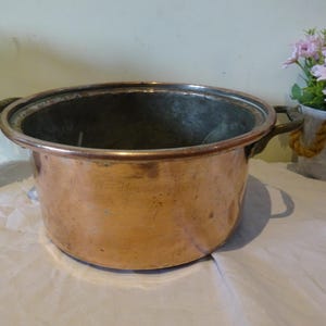 May include: A large copper pot with two handles. The pot is empty and has a shiny, metallic finish.