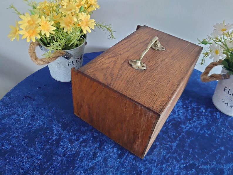 May include: A rectangular wooden box with a brass handle. The box is made of dark brown wood and has a hinged lid. The handle is curved and attached to the top of the box with two screws. The box is sitting on a blue surface.