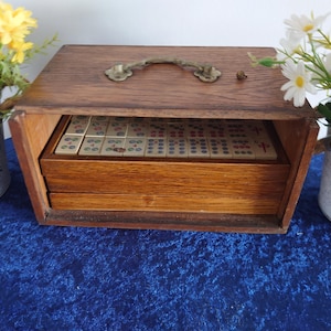 May include: A vintage wooden Mahjong set in a rectangular box with a brass handle. The box is open, revealing the tiles inside a drawer. Yellow and white flowers in decorative pots flank the box on a blue surface.