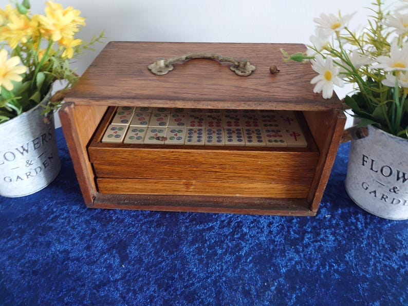 May include: A vintage wooden Mahjong set in a rectangular box with a brass handle. The box is open, revealing the tiles inside a drawer. Yellow and white flowers in decorative pots flank the box on a blue surface.
