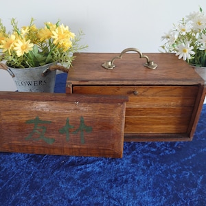 May include: A wooden box with a brass handle and a smaller wooden box in front of it. The smaller box has green text on it. Two small metal buckets with flowers are on either side of the boxes.